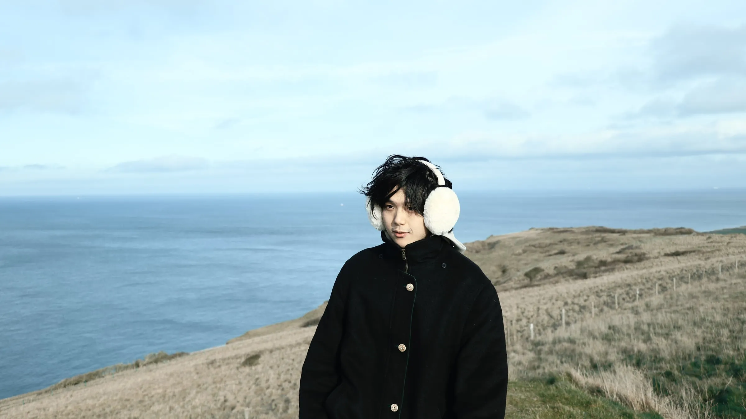 Yichuan (Alan) Lin in a black coat and white earmuffs on a clifftop above the sea