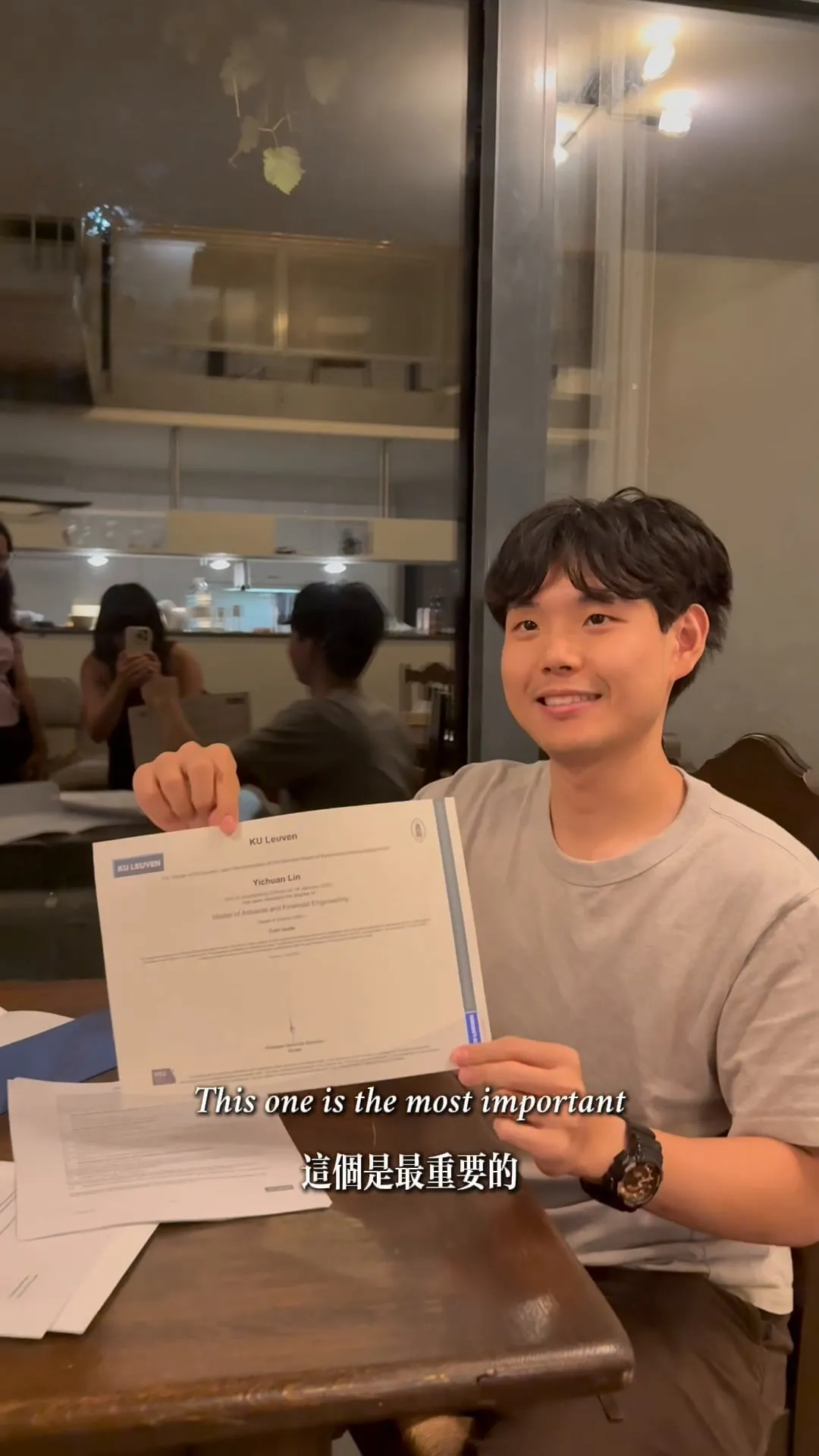 Alan smiling and holding up his KU Leuven diploma at a dining table